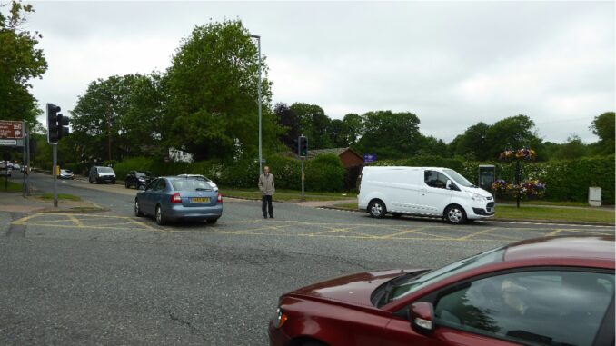 A pedestrian stands in the middle of a junction while the cars go past.