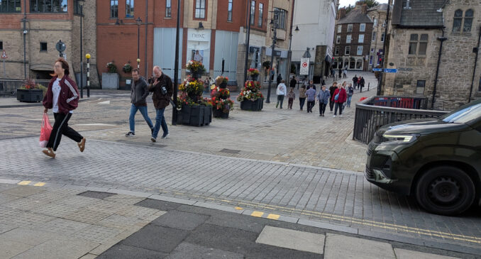 Three pedestrians cross in front of a waiting car. One man holds his hand up to acknowledge the driver.